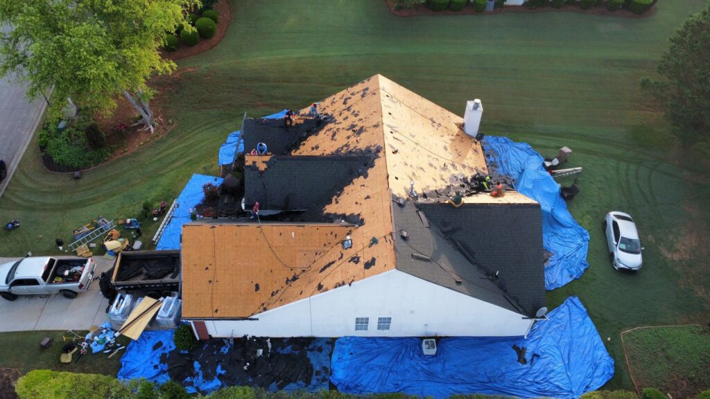 Aerial view of a roof being stripped for repair with blue tarps protecting the surrounding property. 